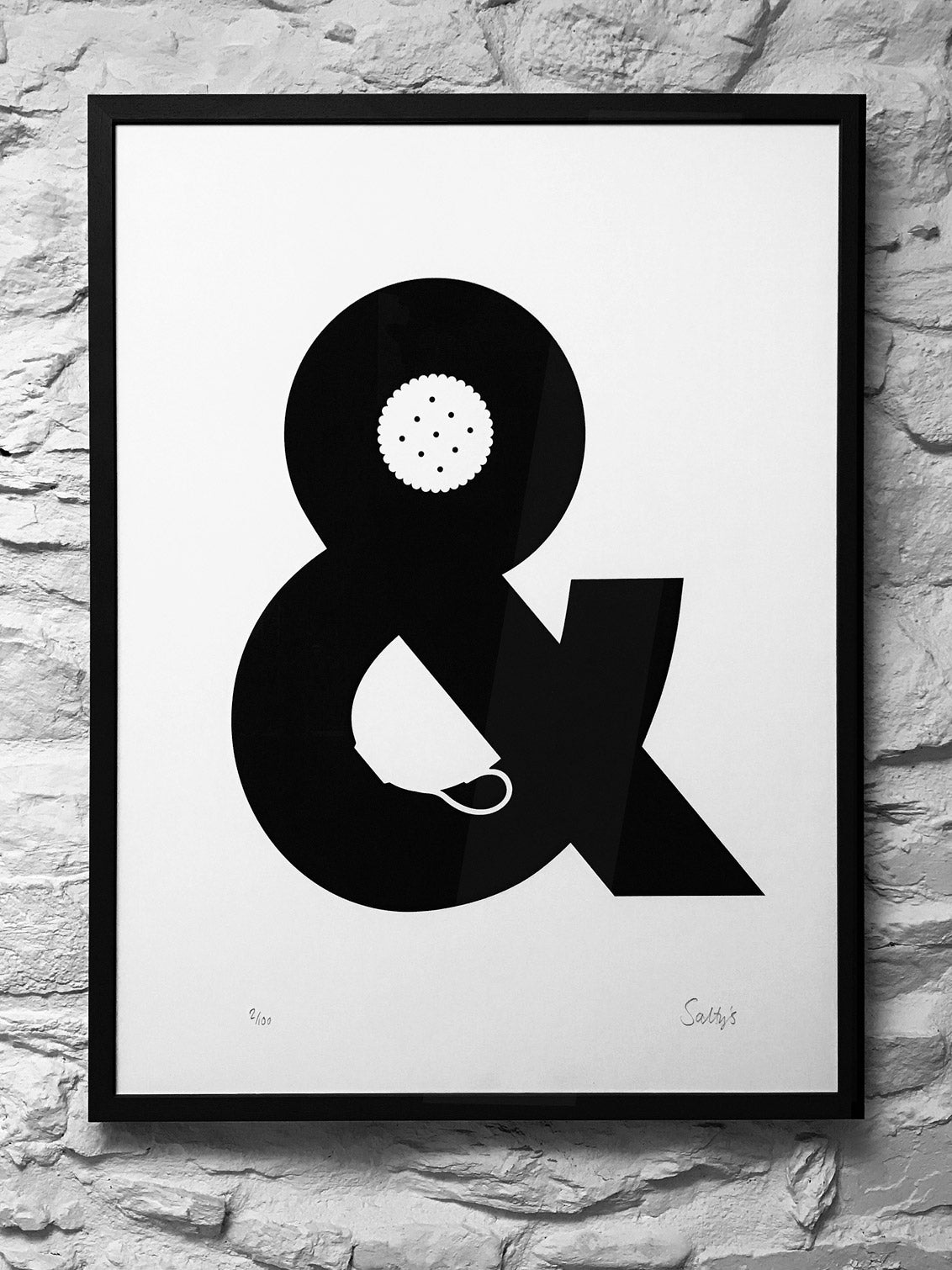 A framed monochrome print hangs on a rough whitewashed Devon wall. The picture is a screenprint of an ampersand with a china cup and a biscuit made out of the negative space. It reads visually as Tea and Biscuits 