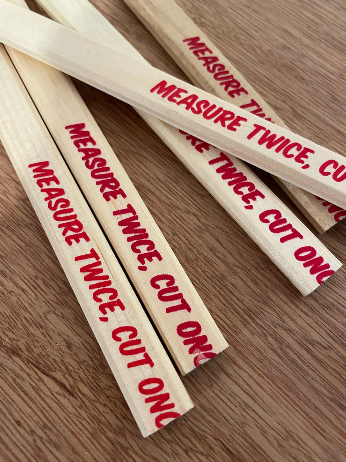 Wooden flat carpenters pencils laid on a wooden table showing the unsharpened end, with the lead within. The pencils have a slogan Measure Twice Cut Once screenprinted in red ink - the joke is that last word has been cut off, or mispositioned. 
