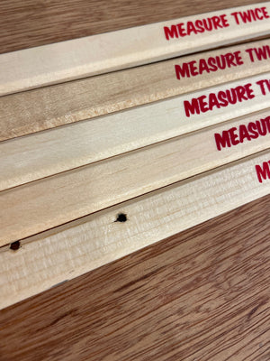 Wooden flat carpenters pencils laid on a wooden table. The pencils have a slogan Measure Twice Cut Once screenprinted at one end in red ink, but the joke is that last word has been cut off, or mispositioned. 