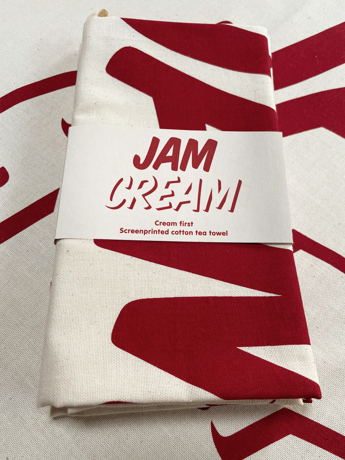A Cream First tea towel folded up with a belly band around, sat on a opened out teatowel of the same design. It says the word Jam, set above the word Cream in a modern bold font, part of the image is seen folded up on the teatowel.