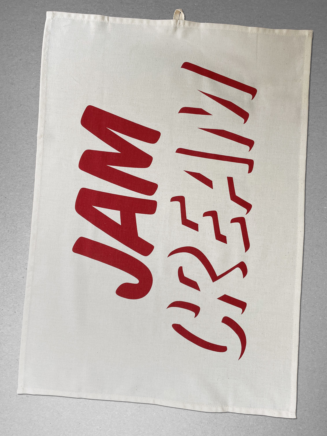 Cleanly printed type in strawberry red ink on a cream coloured organic cotton teatowel, with hanging loop, reading CREAM across the bottom with JAM written on top! A flat lay view from above 