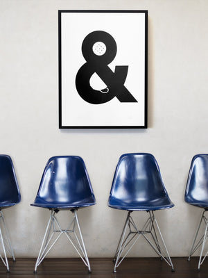 A framed monochrome print hangs on white wall above a row of Eames chairs in blue, The picture is a screenprint of an ampersand with a china cup and a biscuit made out of the negative space. It reads visually as Tea and Biscuits