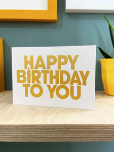 Happy Birthday screen printed in gold ink on a white landscape card, stood on a plywood shelf with hints of framed artwork around the edges , duck egg blue background.