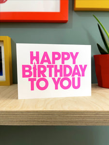 Bright pink text screen printed on a white landscape card, the bold capital font contains a candle created in the negative space of the words. The card sits on a plywood shelf with coloured frames showing at the edges and part of a plant in shot. 