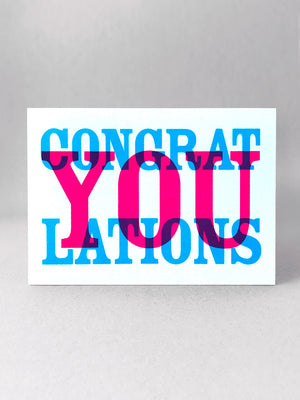This landscape card reads Congrat YOU lations printed in cyan and magenta in overlapping colours. Stood front on, in a light grey studio set.