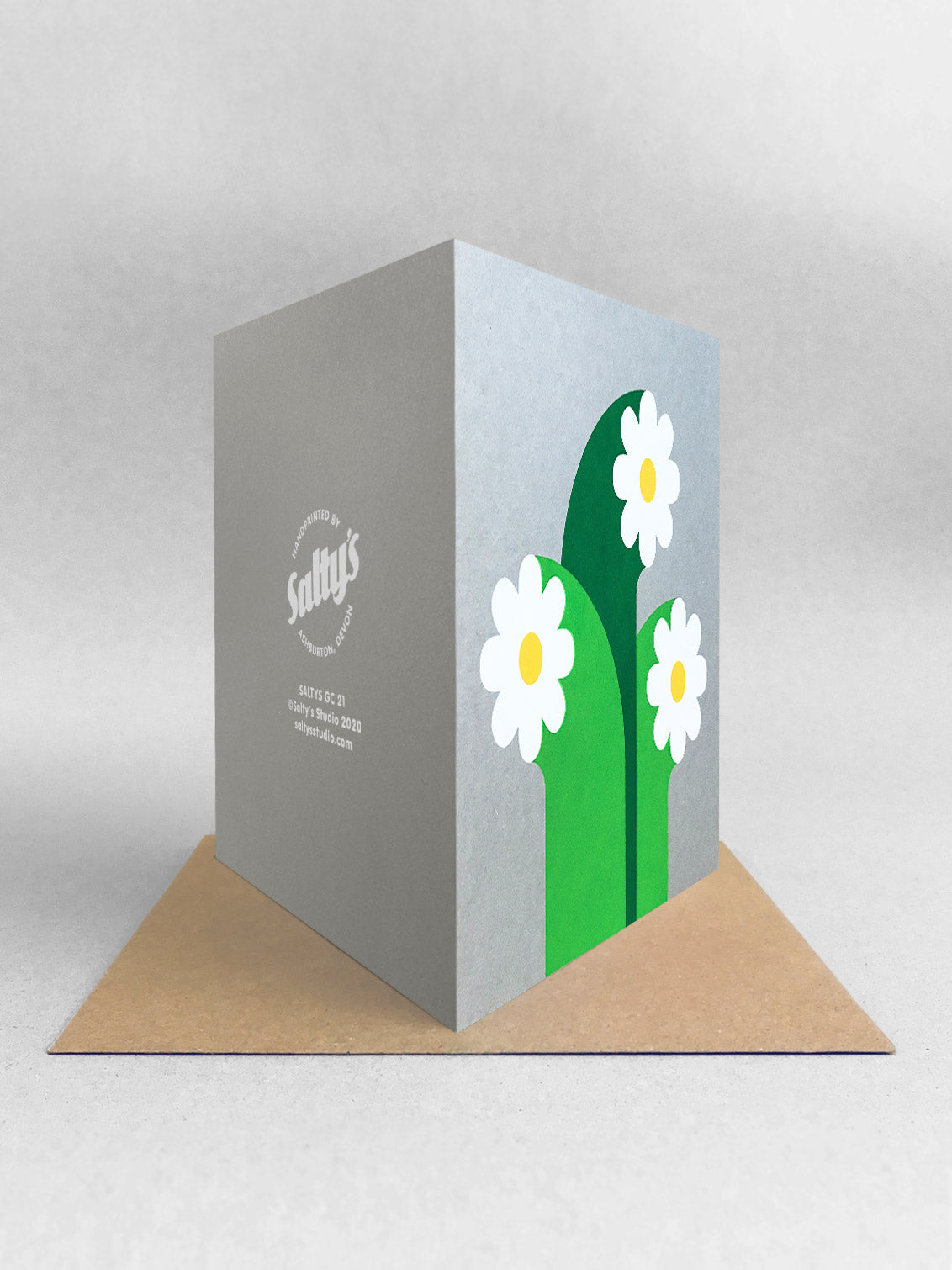  Rear view of the Cosmos card, three white flowers with graphic wide stalks and golden yellow circle centres, screenprinted onto grey card, stood on a brown kraft envelope in a light grey studio shot.