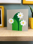 three white flowers with graphic wide stalks, screenprinted on grey card, sit on a ply wood shelf with hints of yellow frames around and a plant showing at the side.
