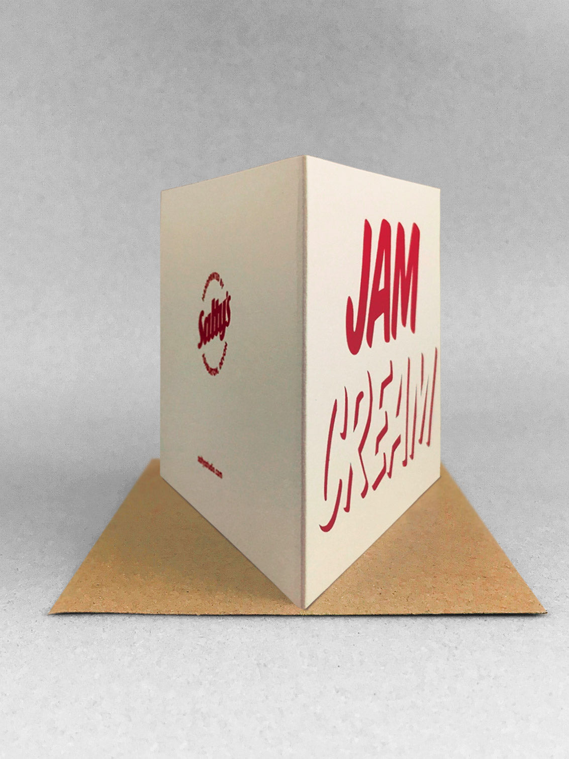 Rear view of cleanly printed type in strawberry red ink on a cream coloured recycled card stock, reading CREAM across the bottom with JAM written on top! Card stood on a kraft envelope in a light grey studio.