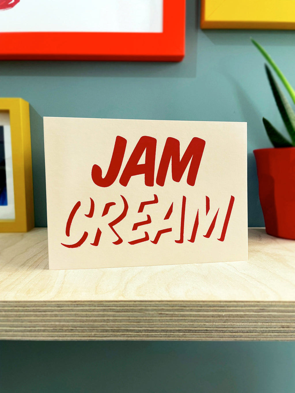   Cleanly printed type in strawberry red ink on a cream coloured recycled card stock, reading CREAM across the bottom with JAM written on top! Card stood on a plywood shelf with coloured pictures frames showing around the edges and a plant to one side.