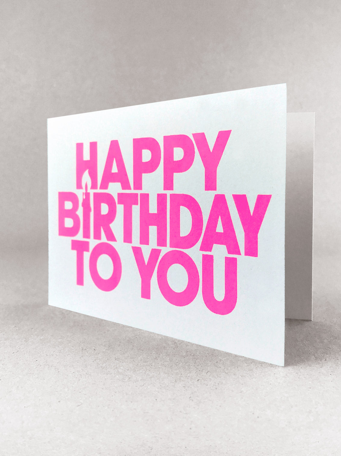 Happy Birthday to You landscape greetings card. The I of Birthday is a candle using negative space. A ¾ view to show the plain inner. Printed in capital letters, simple font printed in neon pink ink on a white card made by Salty's Studio.