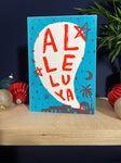  Al-le-lu-ya handprinted Christmas card in turquoise and red, sat on a plywood shelf with baubles and greenery about.
