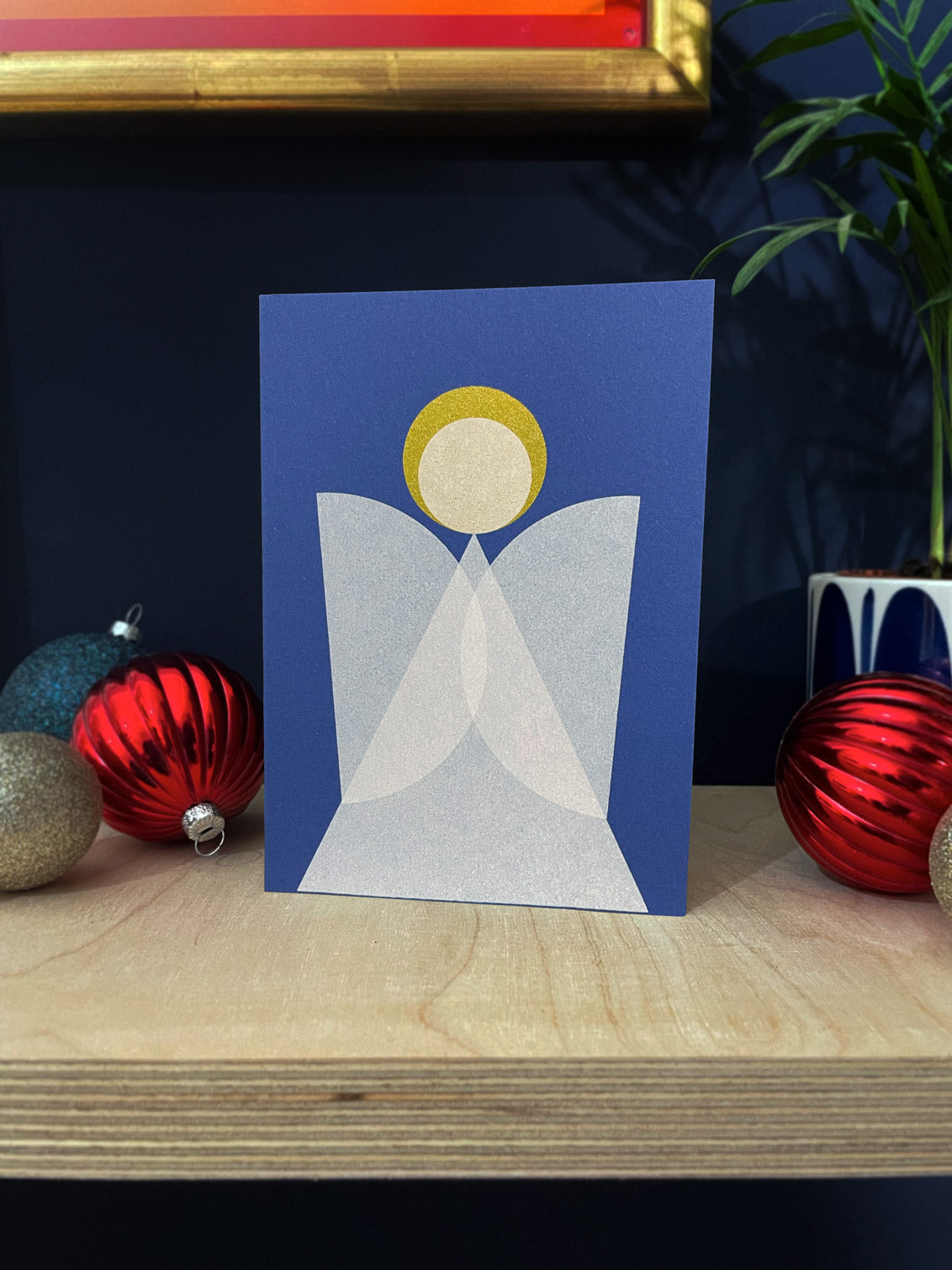 Geometric angel design, blue card, white overlapping wings and a gold halo. Sits on a plywood shelf with baubles and foliage about.