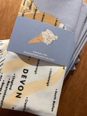 A pile of folded teatowels in wraps lie on a wooden surface. The Devon and Cornwall Ice Cream Map design is howling both on the label and showing on the teatowel itself. 