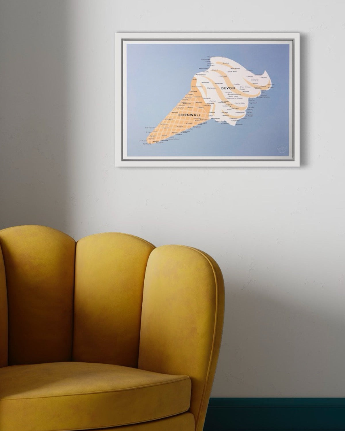 A framed print of an ice cream that also happens to be in the shape of Devon and Cornwall, in the South West of England. Is on a white wall above a curvy yellow velvet chair, with a scalloped back. There are soft shadows.