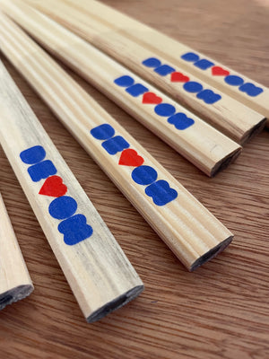 Carpenters pencils lying on a wooden table top, the flat end shows a glimpse of the unsharpened lead. The printed word of Devon, with the V as a heart and in royal blue and red is shown across all of them.