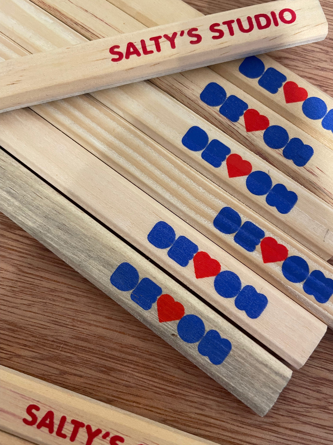 Carpenters pencils lying on a wooden table top. The printed word of Devon, with the V as a heart and in royal blue and red is shown across all of them. The reverse is printed in red saying Salty's Studio