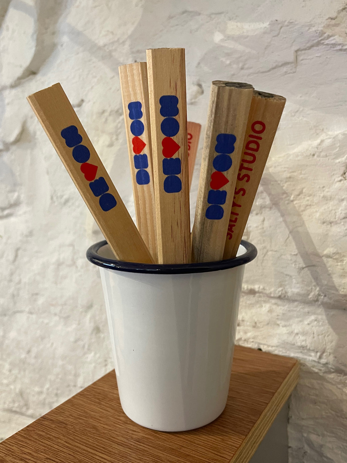 several wooden flat carpenters pencils are stood in a white enamel pot on a plywood shelf. One end of the pencils has been printed with Devon in blue and red, with the V as a heart. Stood against a whitewashed rough stone Devon wall