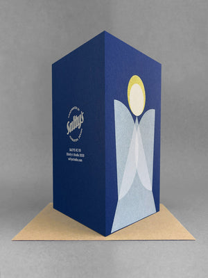 Rear view of a geometric angel christmas card design, on blue card, white overlapping wings and a gold halo. Stood on a brown envelope in a light grey studio.