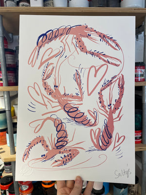 Hand holds up a print of four pink cartoon langoustine, who are flung into a frenzied swirl, with hearts and scribbles making this a joyful scene, screenprinted in pink with blue accents. Held up against a backdrop of colourful screenprinting inks. 