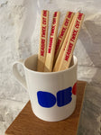 Wooden flat carpenters pencils sit in a china mug on a plywood shelf against a whitewashed wall. The pencils have a slogan Measure Twice Cut Once screenprinted at one end in red ink, but the joke is that last word has been cut off, or mispositioned. 