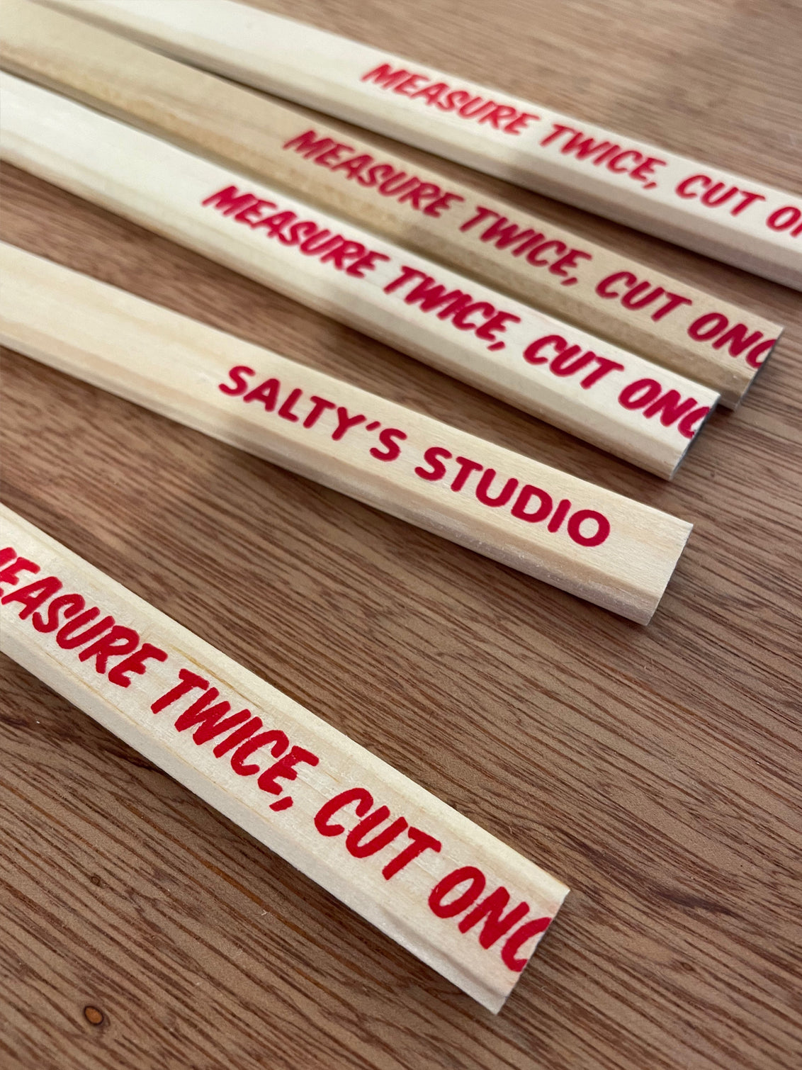 Wooden flat carpenters pencils laid on a wooden table. The pencils have a slogan Measure Twice Cut Once screenprinted at one end in red ink - the joke is that last word has been cut off, or mispositioned. The rear reads Salty's Studio