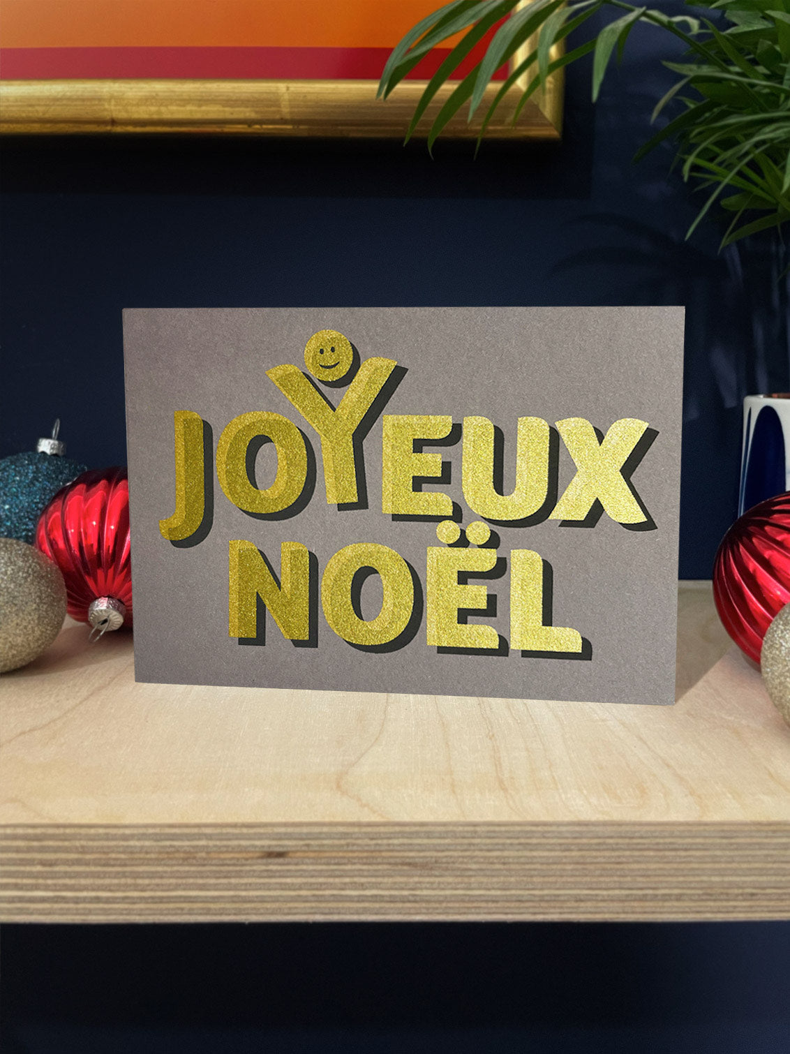 Golden text screenprinted onto grey card stock, spells out Joyeux Noel with a happy face above the Y, as though celebrating. Landscape card stood on a plywood shelf with baubles around against a dark blue wall.