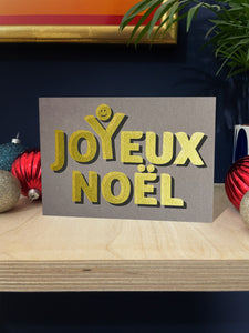 Golden text screenprinted onto grey card stock, spells out Joyeux Noel with a happy face above the Y, as though celebrating. Landscape card stood on a plywood shelf with baubles around against a dark blue wall.