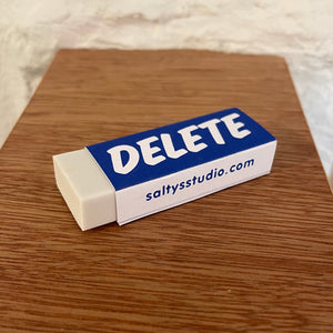 A close up, slight angled photo of a white eraser, it has a blue paper wrap around it with the words DELETE written upon and then on the side is saltysstudio.com The eraser is on a wooden ledge against a white wall slightly blurred in the background.