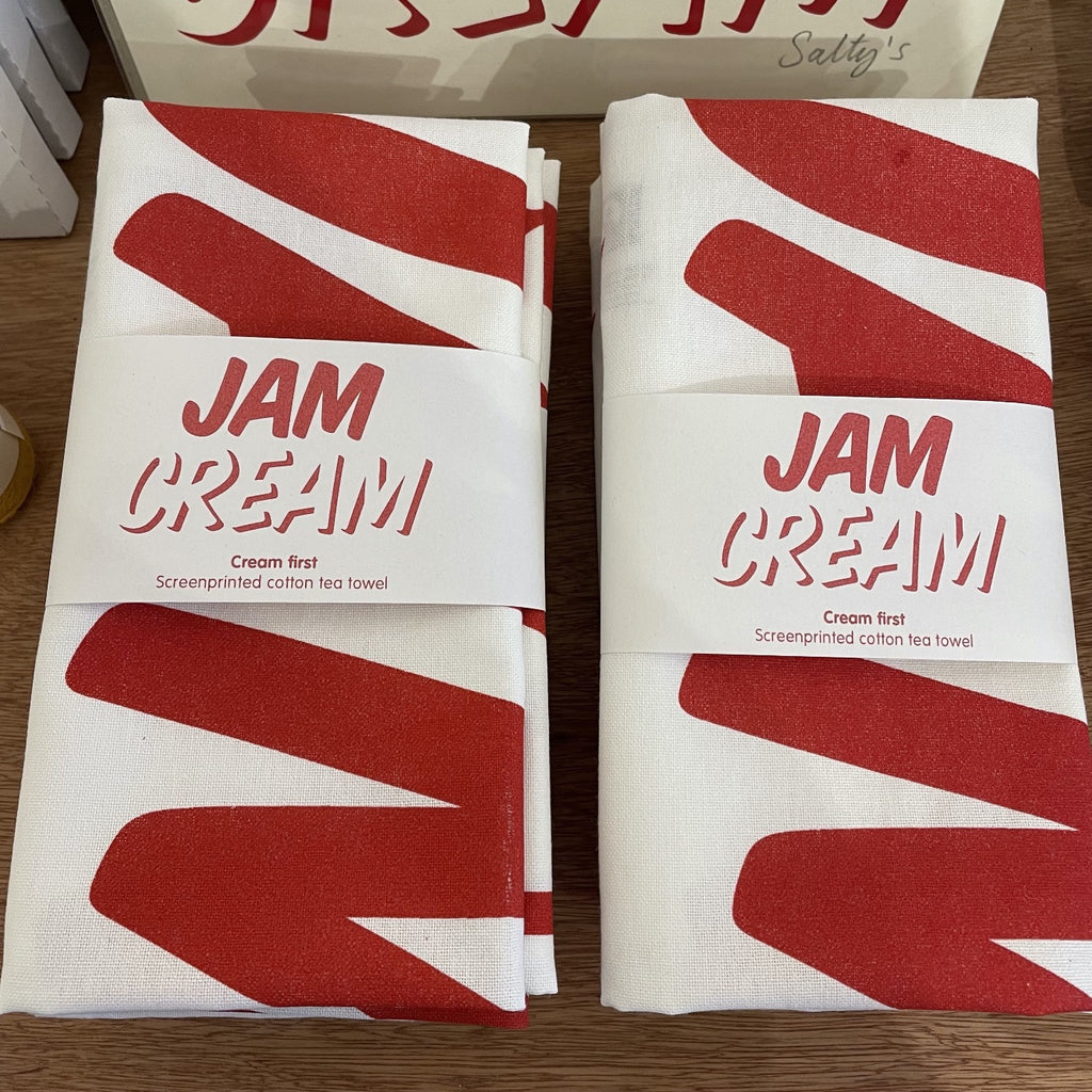 Two Cream First tea towels folded up side by side with a belly bands around, sat in piles on a wooden table top. It says the word Jam, set above the word Cream in a modern bold font, part of the image is seen folded up on the teatowel.