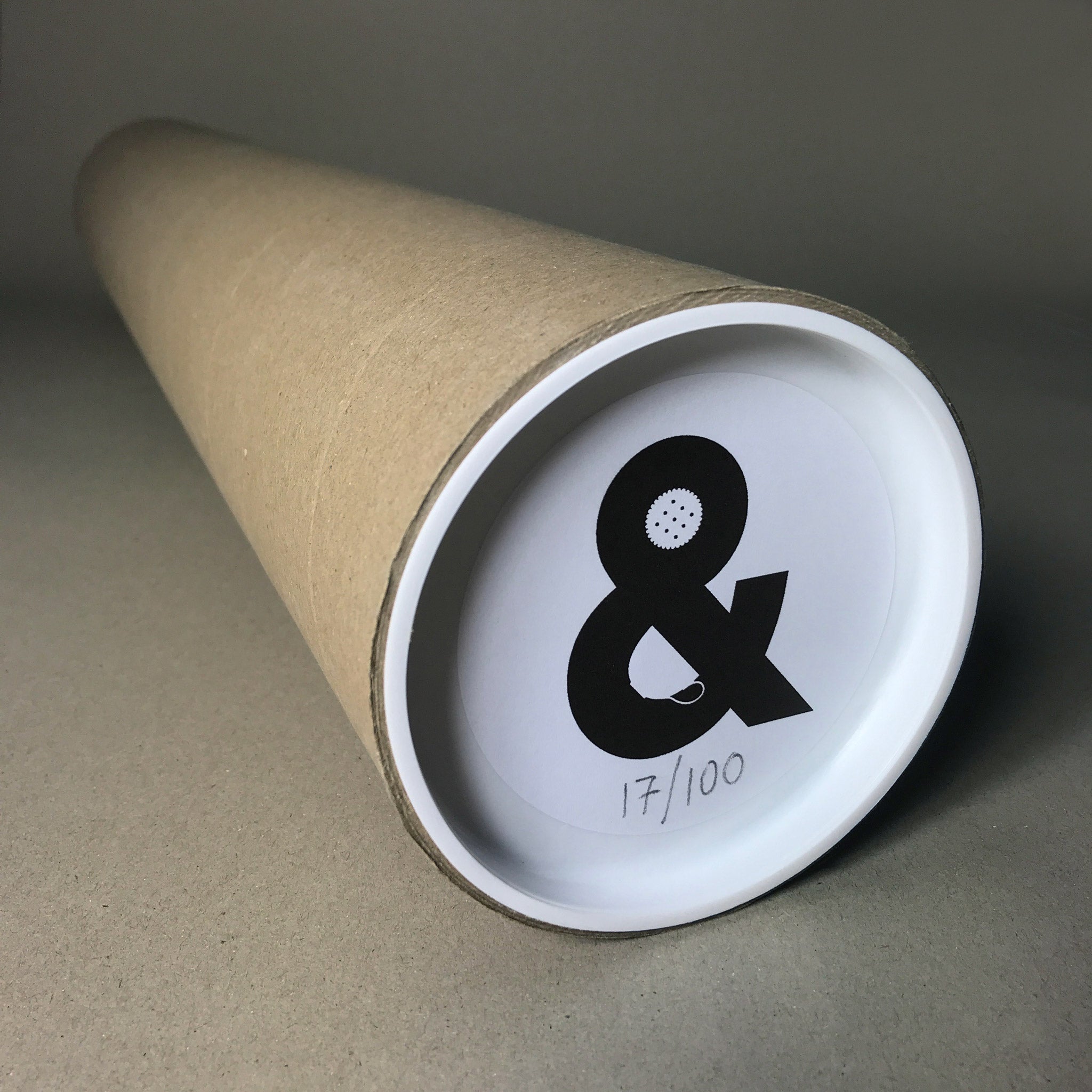 A cardboard tube laid on a plain grey background, containing the tea and biscuits ampersand with a sticker on the end depicting that.